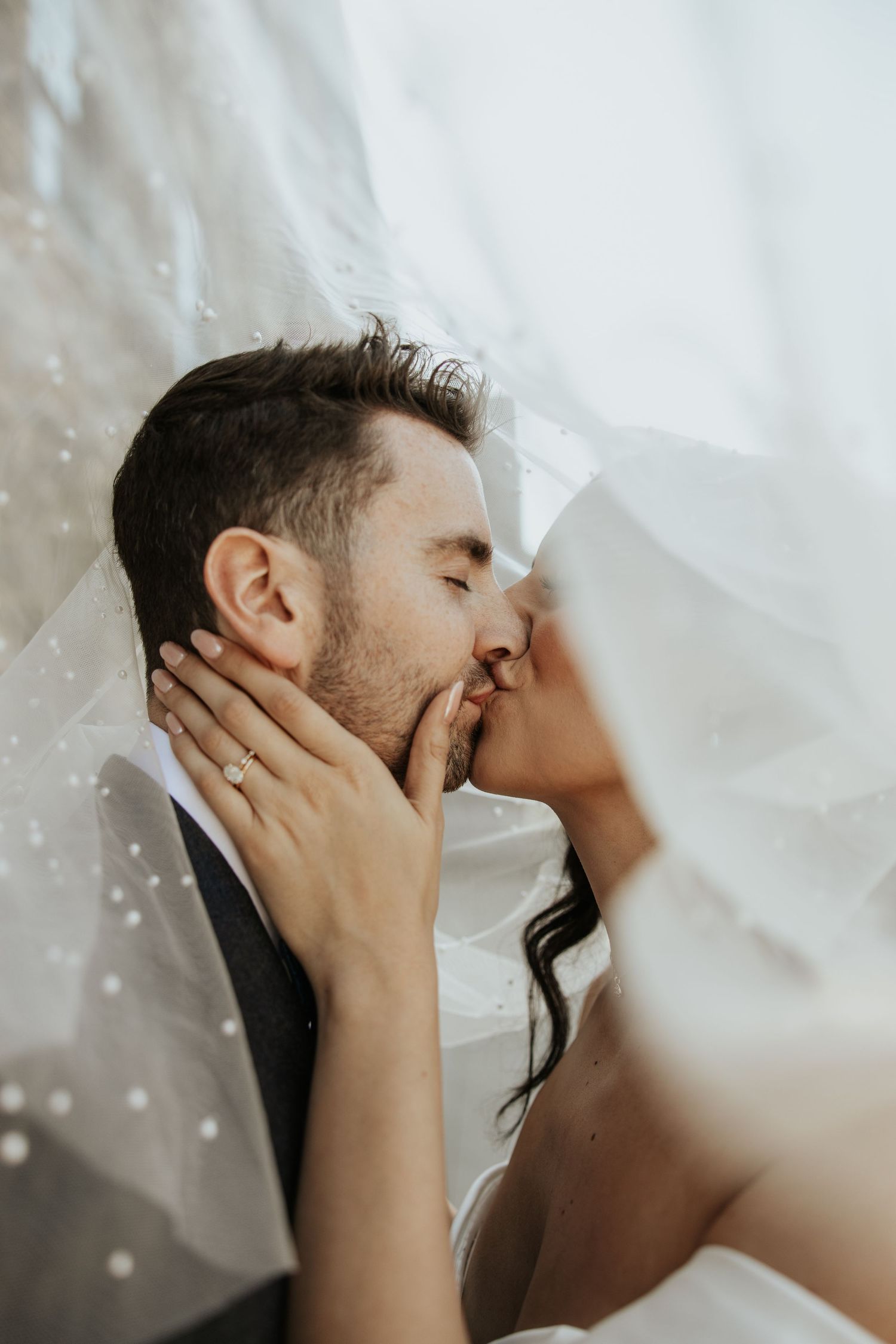 Romantic close-up of newlyweds kissing under flowing white veil.