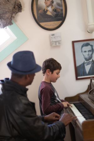 A piano lesson in progress with an instructor in a navy hat and student in a burgundy shirt.