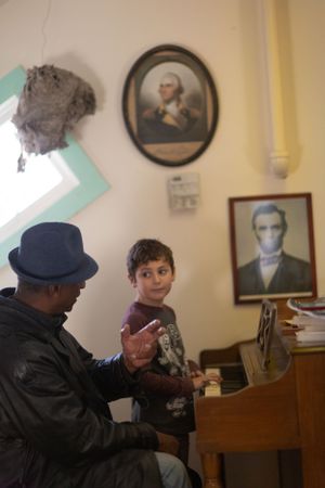 Two people practicing music together at a piano in a room with framed portraits on the wall.