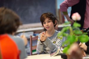 Students sitting at desks in a classroom with a chalkboard and plants visible.
