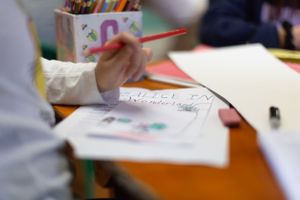 Close-up of hands writing with red pencil on paper in classroom setting.