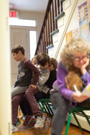 Students seated on green chairs in a classroom with wooden staircase visible.