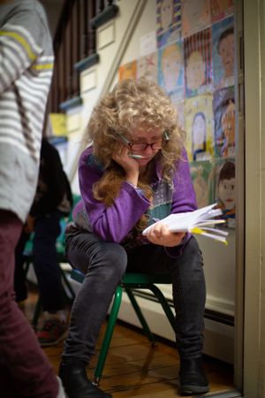 Student working intently while seated on a green chair in classroom corner.