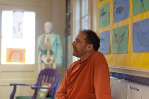 Person in orange sweater sitting in classroom with colorful artwork and anatomical model visible.