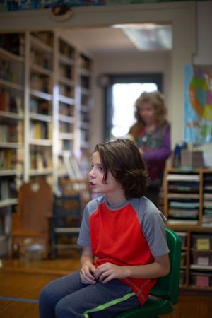 Student in red vest sitting attentively in classroom with bookshelves.