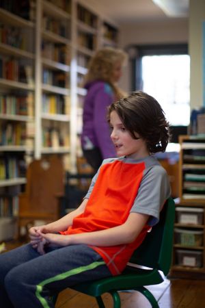 Student in red vest seated in classroom with bookshelves and educational materials.