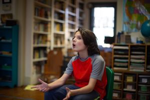 Student in red and grey speaking animatedly while seated in classroom.
