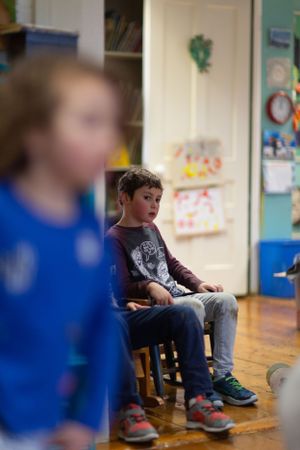 Children sitting in chairs along a classroom wall decorated with artwork.