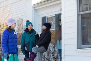 Group of people in winter coats gathered outside a white building in the snow.