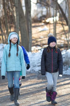 Two children in winter coats walk along a snowy trail through bare trees on a winter day.