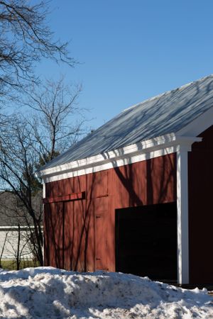 A red barn with white trim and snow on the ground casts tree shadows against its walls on a sunny winter day.