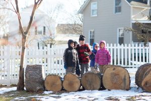 Children stand on stacked logs in a snowy yard with a white picket fence in the background.