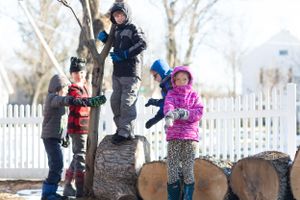 Children playing on stacked logs in a winter yard with white picket fence.