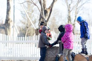 Children playing on snow-covered logs in a winter yard with a white picket fence in the background.