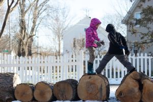 Children balancing and walking across large logs in the snow with a white fence behind them.