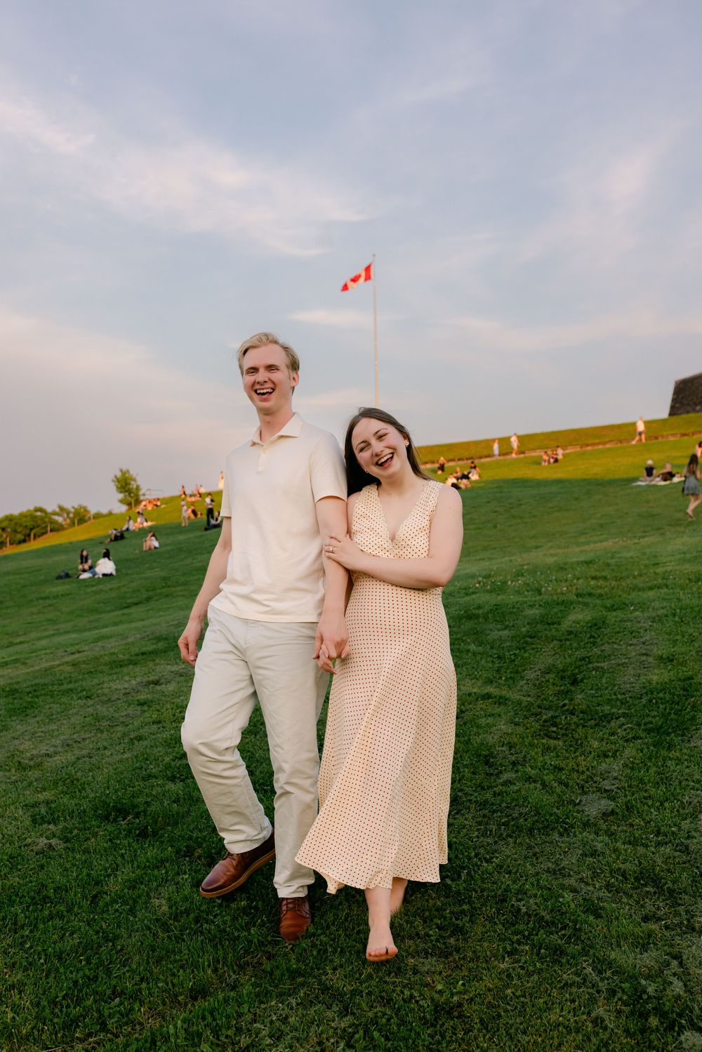 A smiling couple holds hands on a green lawn with a Canadian flag in the background under a cloudy sky.