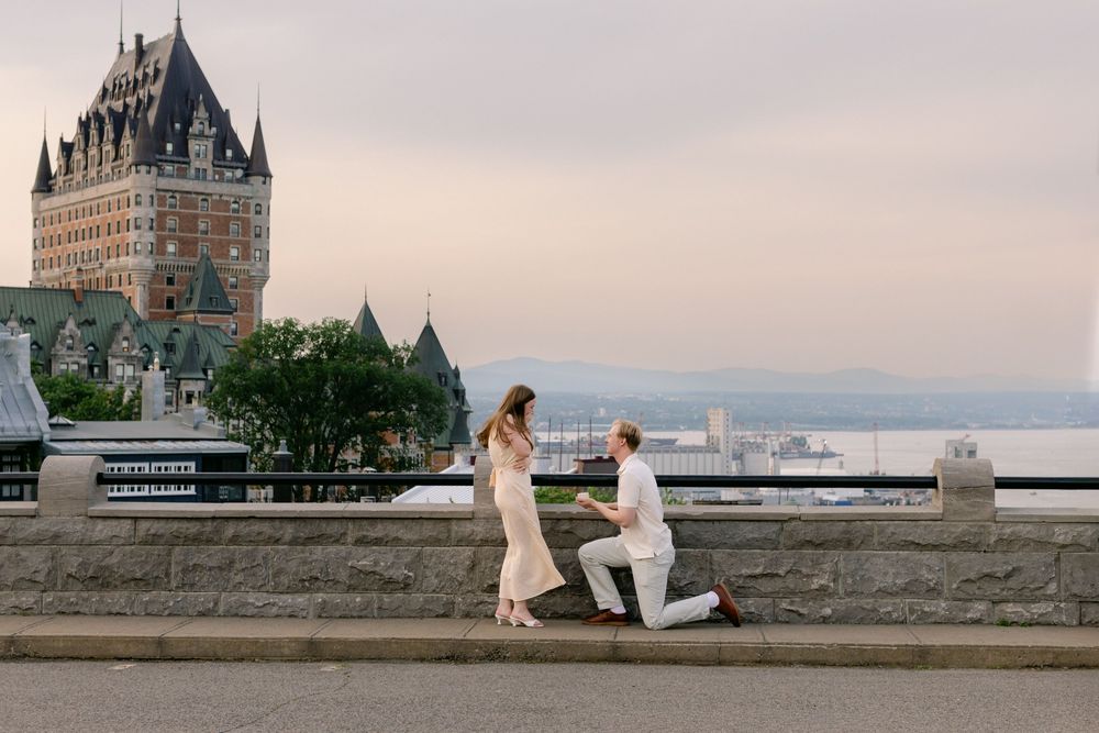 Man proposes on one knee to woman near stone wall with Chateau Frontenac and St. Lawrence River backdrop.