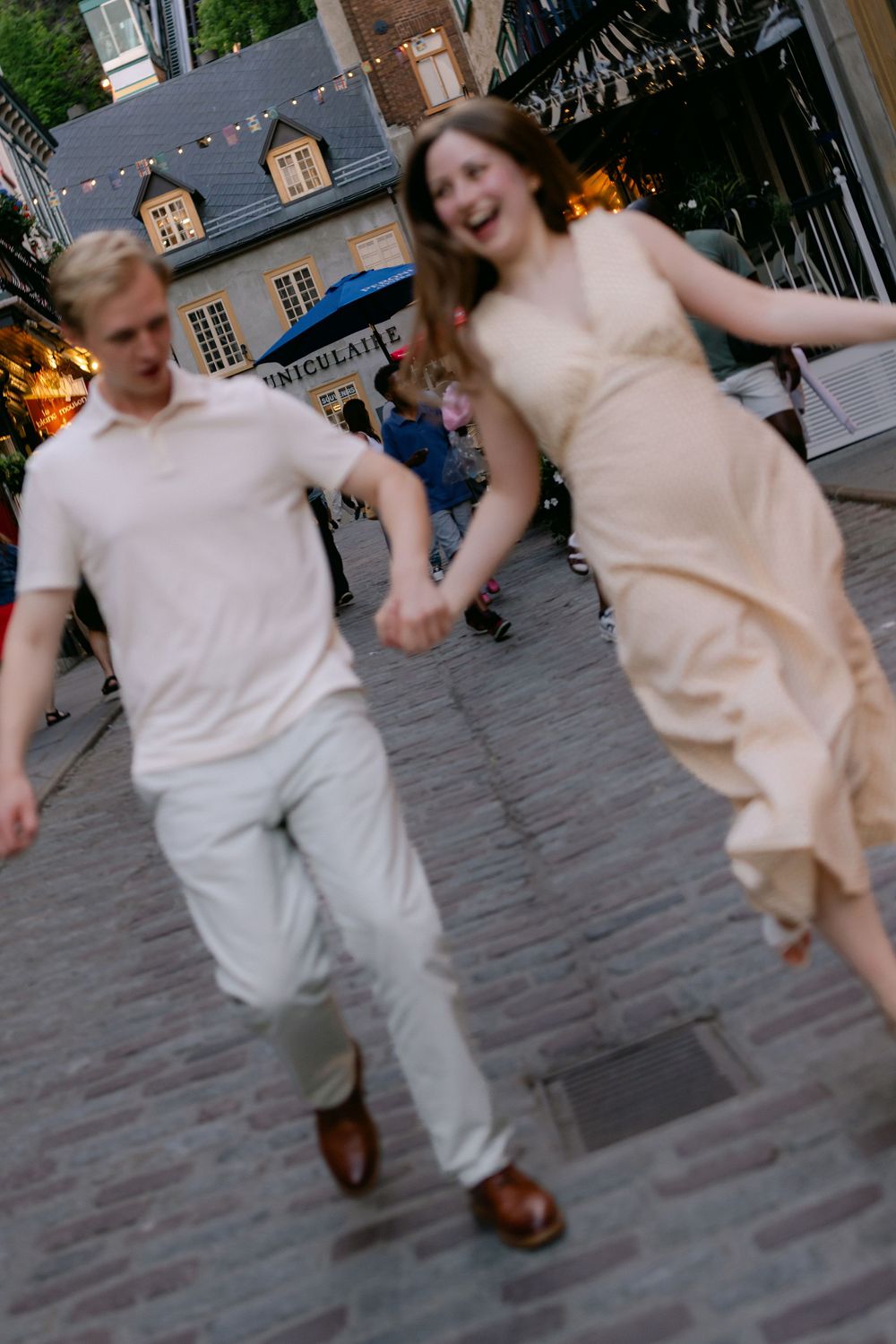 A joyful couple holds hands and runs together on a cobblestone street, the woman in a flowing cream dress laughing.