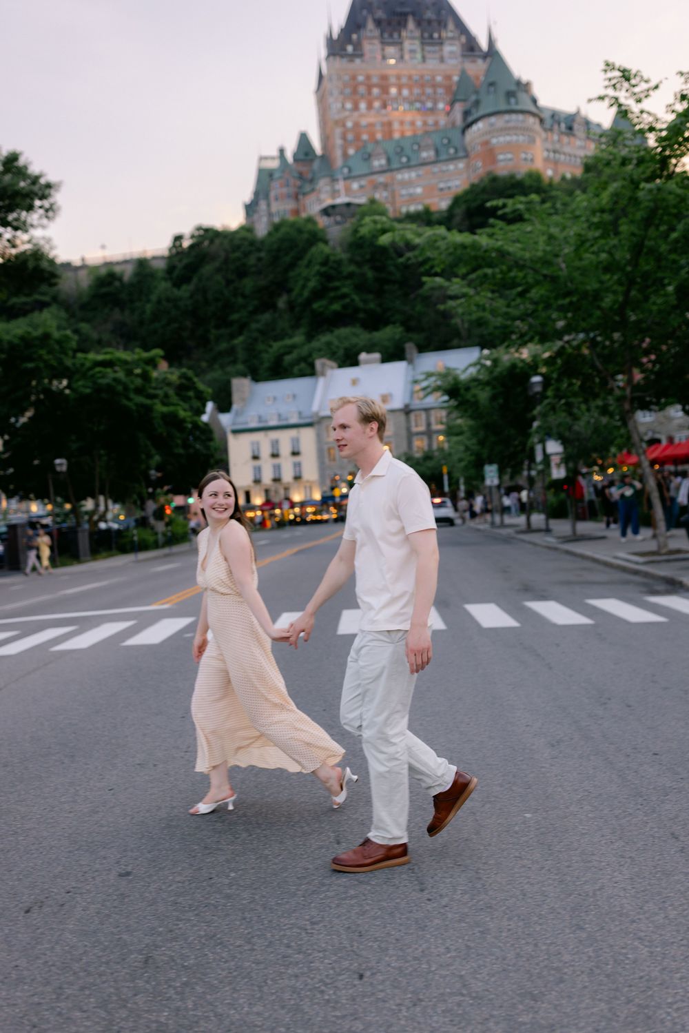 Couple holding hands crossing street in Quebec City with Chateau Frontenac castle in background at dusk.