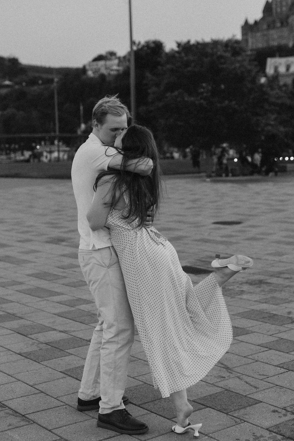 Black and white photo of a couple embracing and kissing in an outdoor plaza, woman lifting her leg romantically.