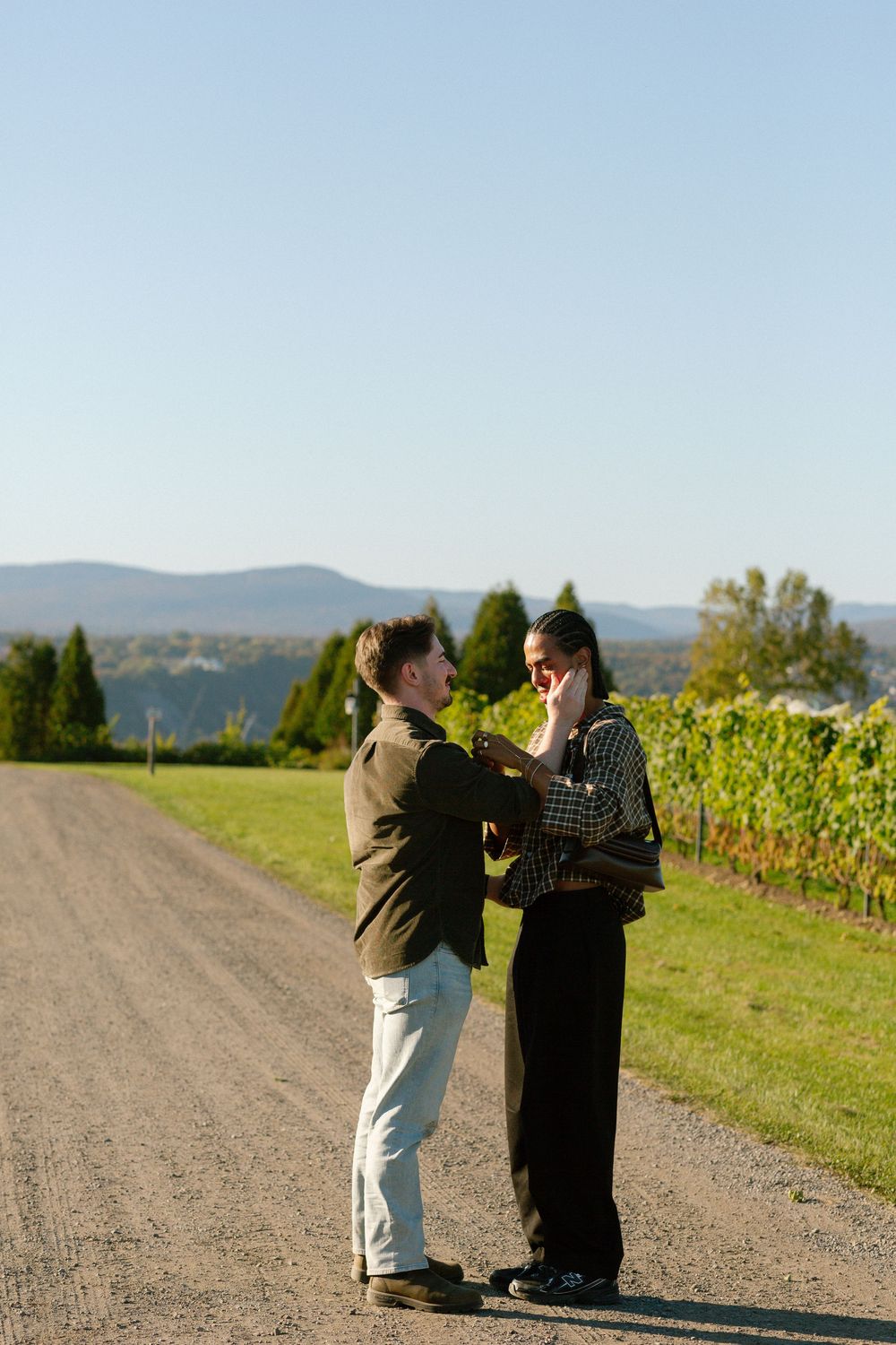 Couple sharing a romantic moment on a scenic vineyard gravel path with mountain views and clear blue sky.
