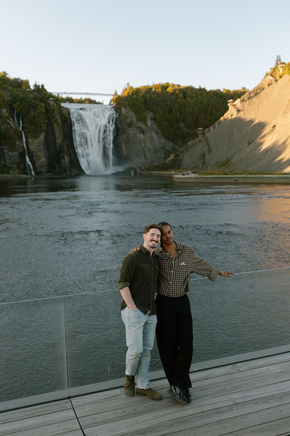 Two friends smiling together on a deck with a stunning waterfall and rocky cliffs in the background.