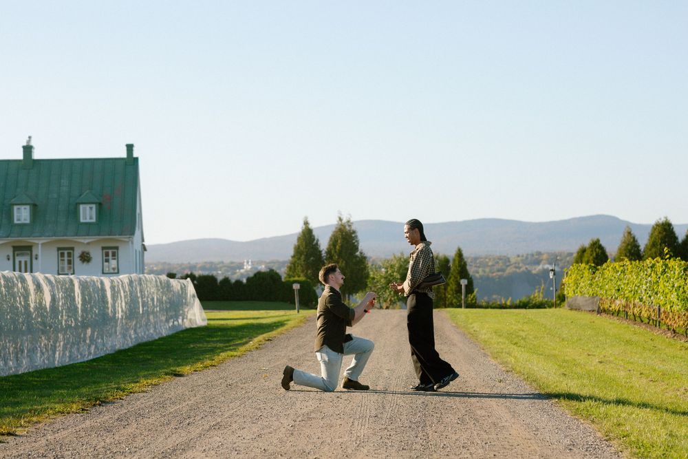 Person proposing on one knee on a gravel path beside a vineyard and white farmhouse with mountain views.