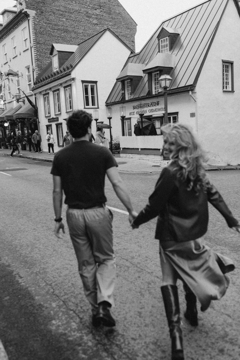 Black and white photo of a couple holding hands while walking across a street in a charming European-style town.