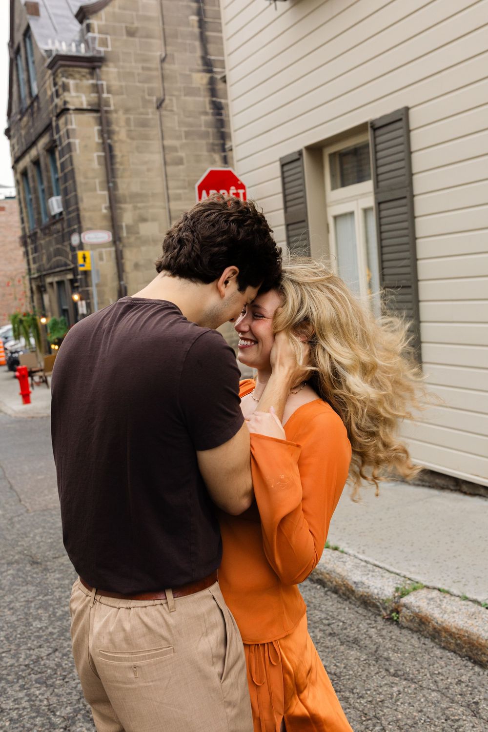 A joyful couple embraces and laughs together on a charming urban street, with the woman wearing an orange dress.