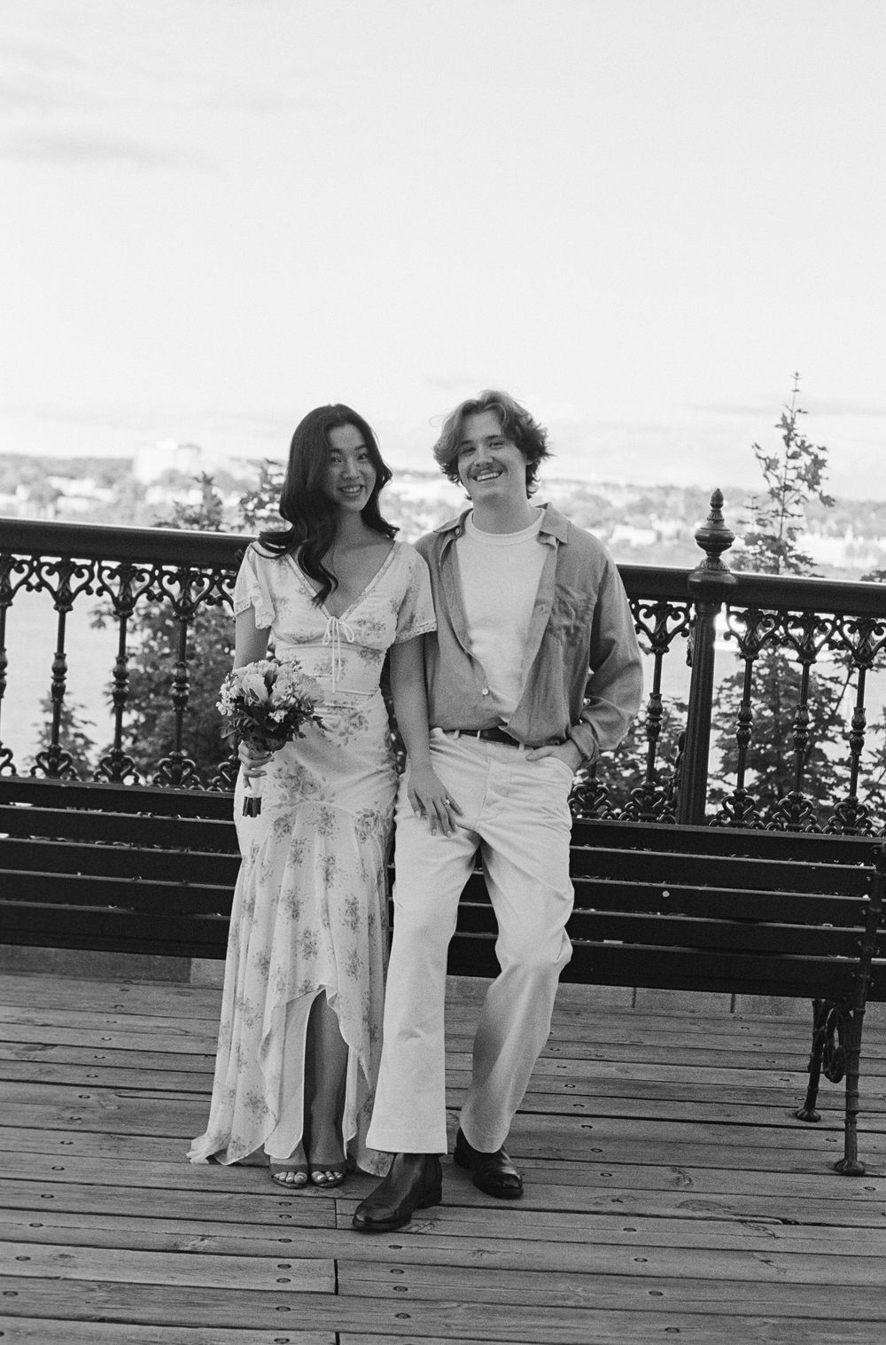 Black and white photo of a smiling couple posing on a wooden deck, woman in floral dress holding bouquet, man in casual outfit.