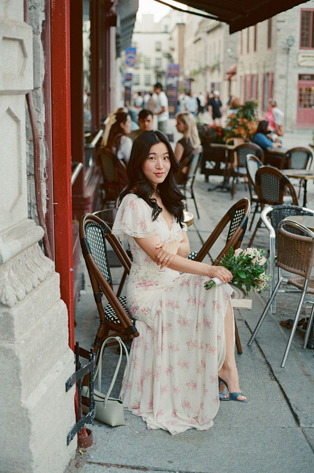 Young woman in floral dress holding flowers, seated at outdoor café patio on a sunny day.