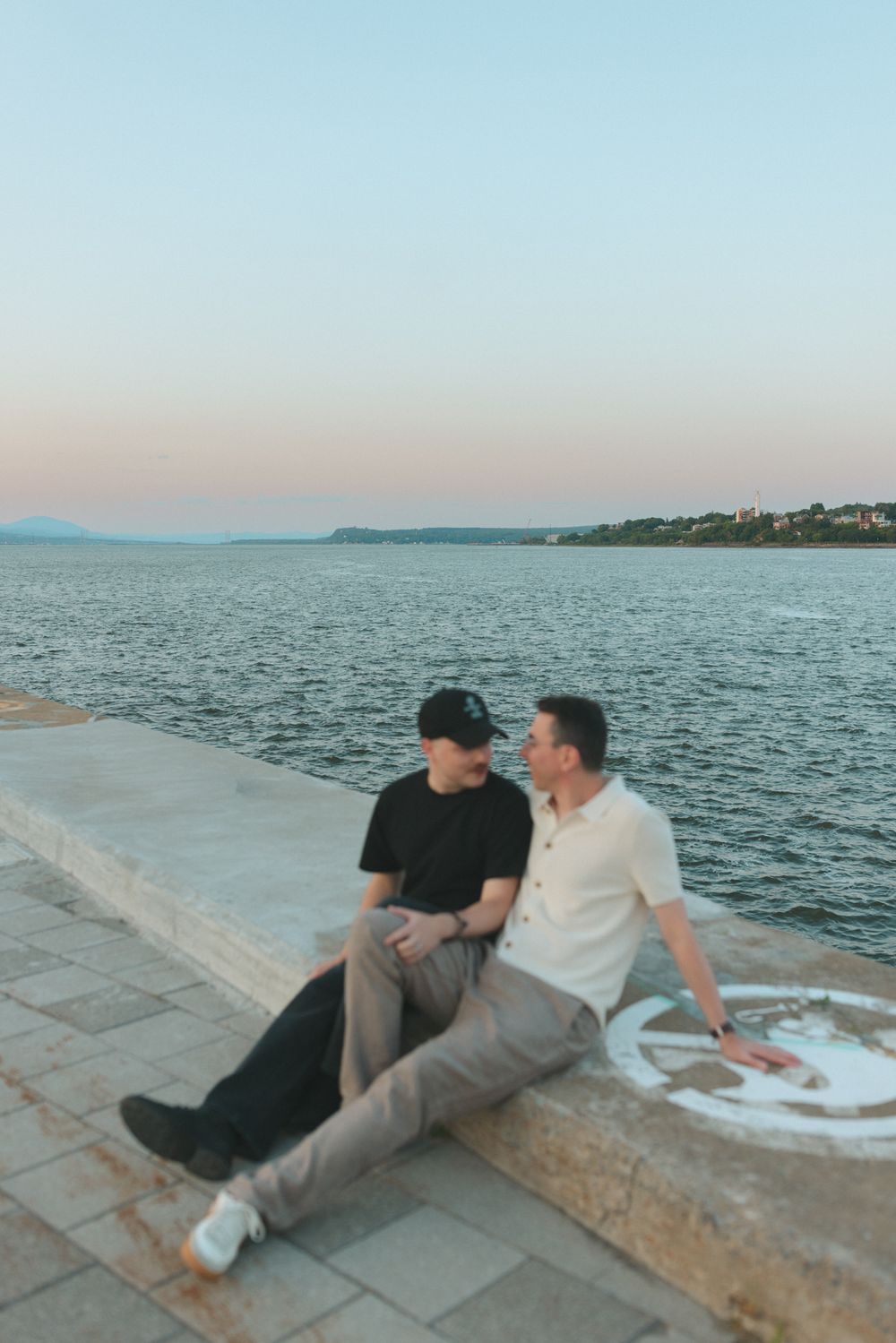 Two men sitting together on a seaside promenade at sunset, sharing a romantic moment by the calm blue water.