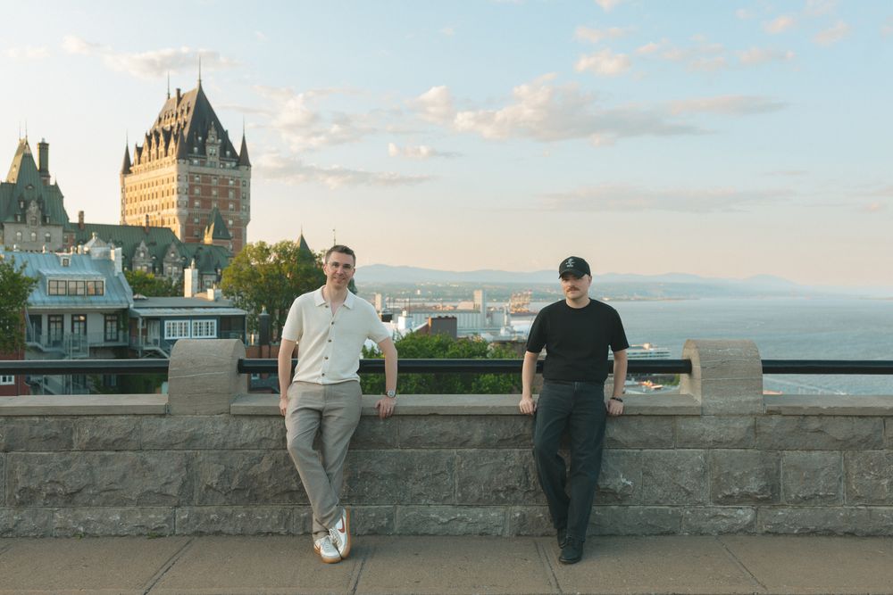 Two men pose by a stone wall with Château Frontenac and St. Lawrence River in Quebec City at sunset.