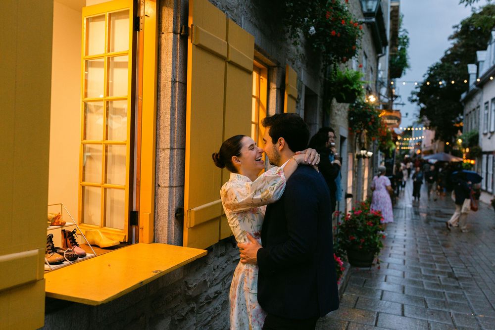 Couple sharing a romantic kiss on a rainy cobblestone street in Old Quebec City at dusk near a yellow building.