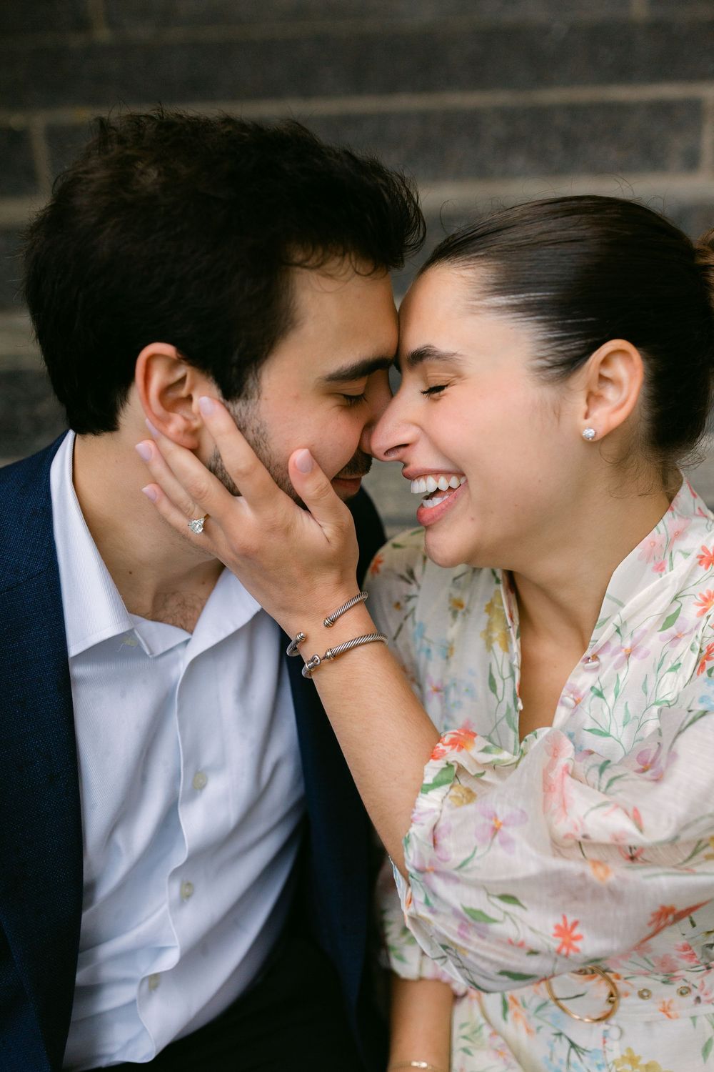 A joyful couple shares an intimate moment, the woman laughing as she touches the mans face, wearing a floral dress and engagement ring.