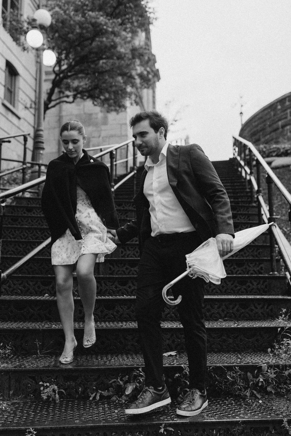 Black and white photo of a couple walking down outdoor stairs, man holding umbrella, woman in dress and jacket.
