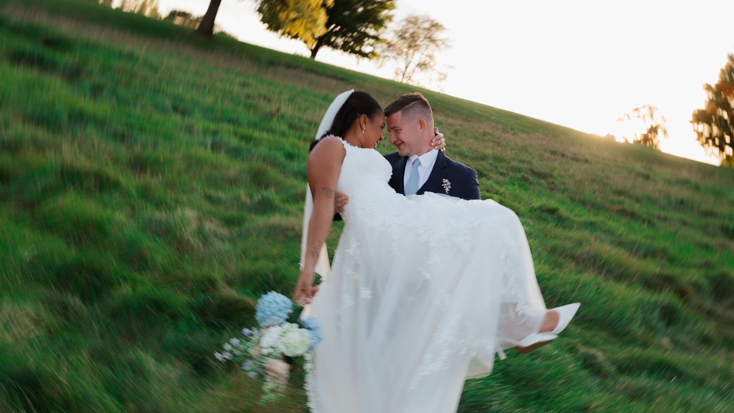 Groom in a navy suit carries his smiling bride in a white gown across a sunlit grassy hillside holding a pastel bouquet.