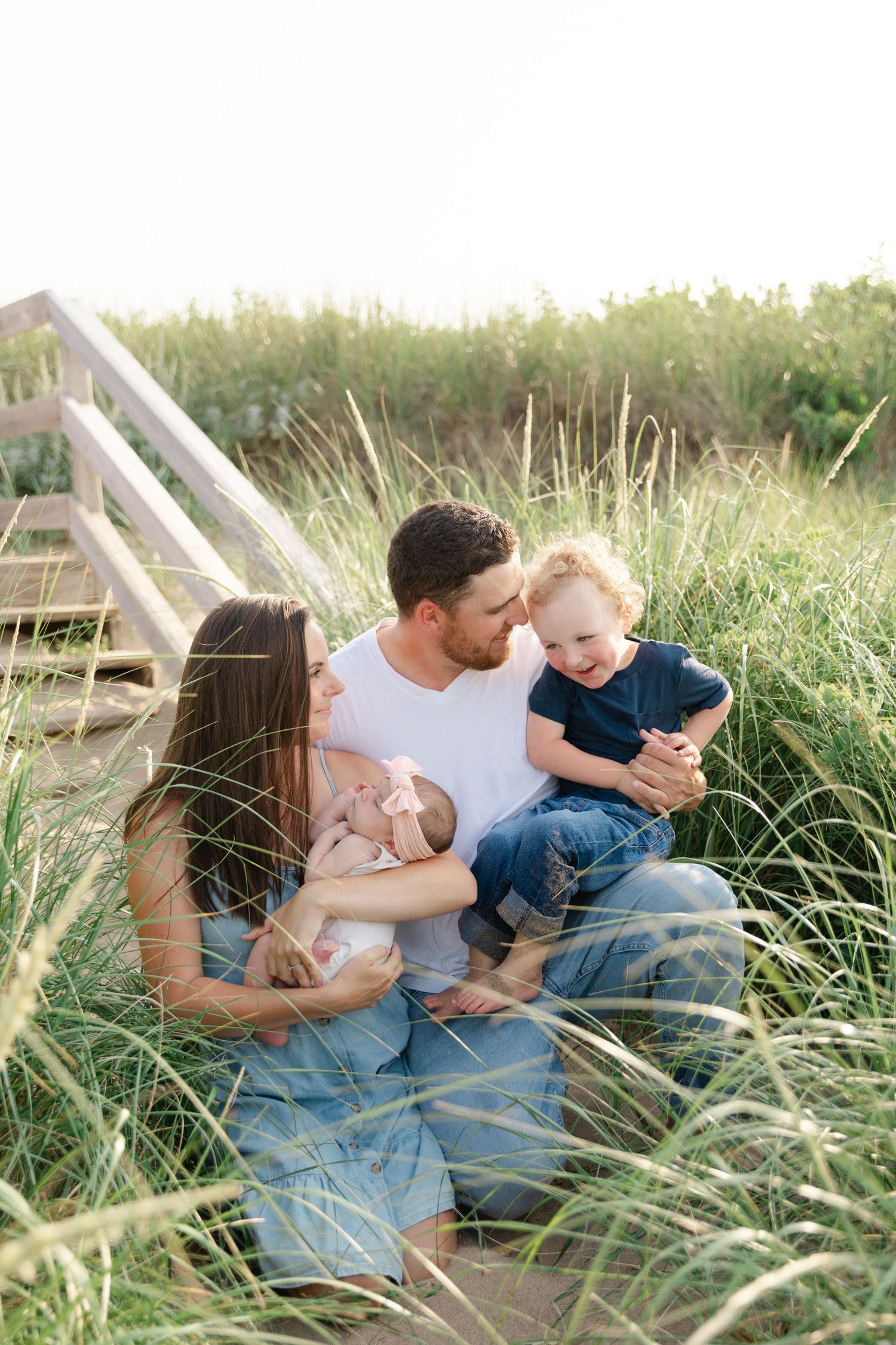 A family sits together on wooden steps surrounded by tall grass during a golden hour photoshoot outdoors.