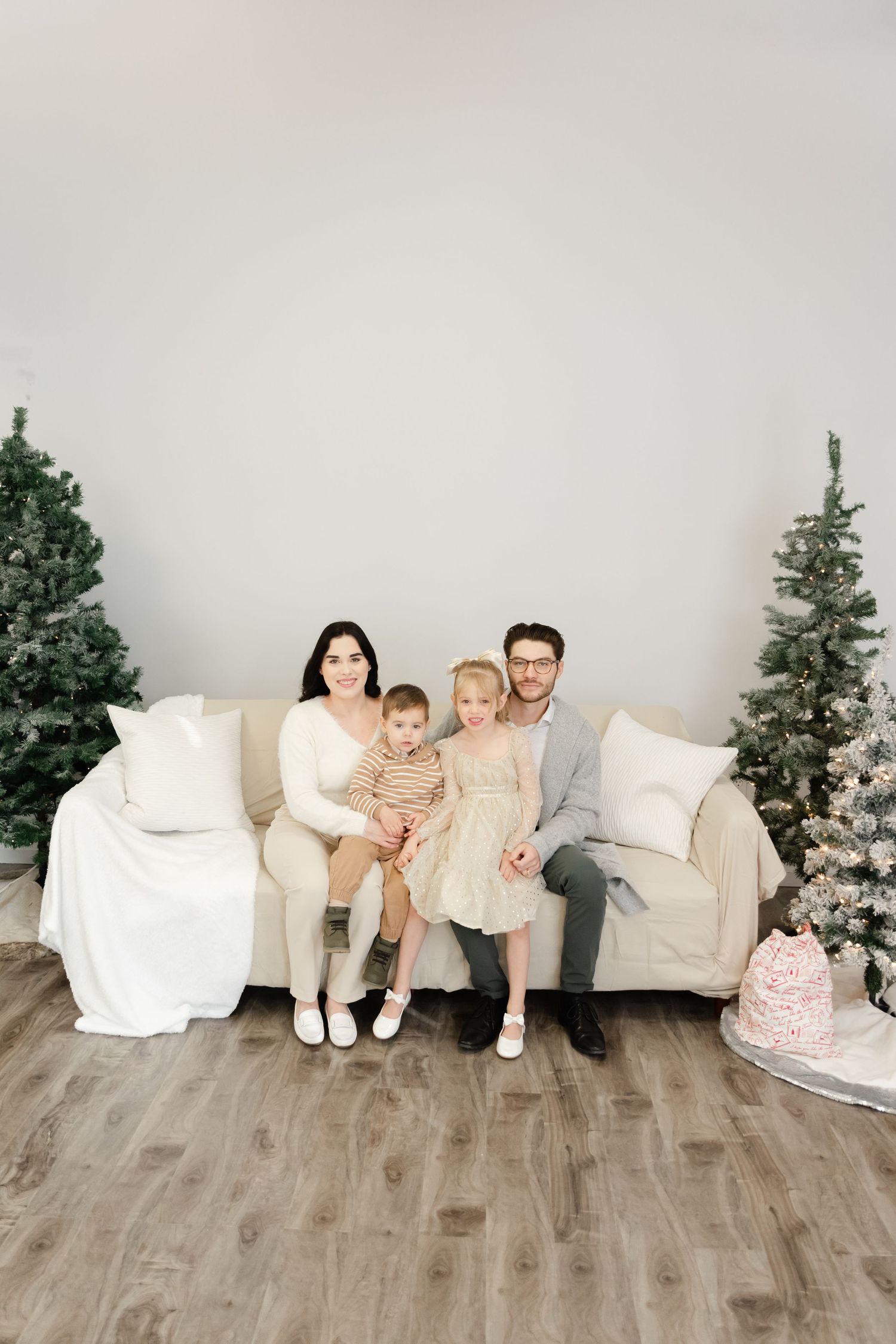 Family sits together on white couch between Christmas trees in minimalist studio setting with wood floors.