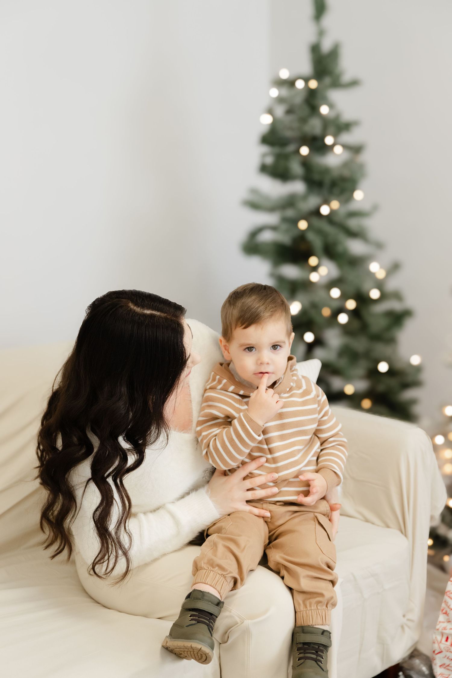 A child in a striped sweater sits with a parent on a cream couch in front of a decorated Christmas tree.