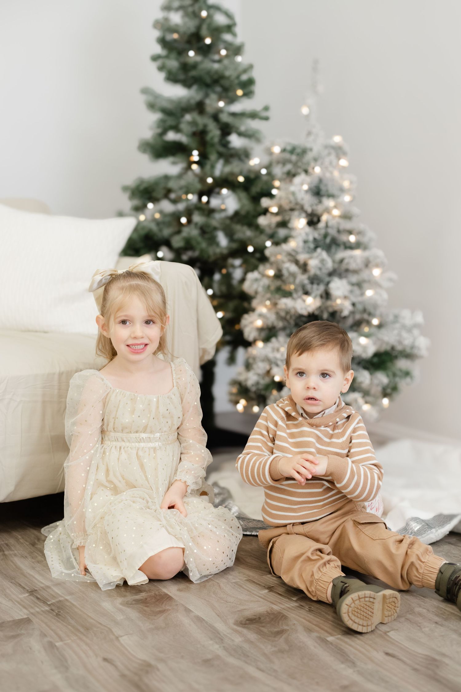 Two young children in holiday outfits sitting on floor by decorated Christmas trees in white room with hardwood flooring.