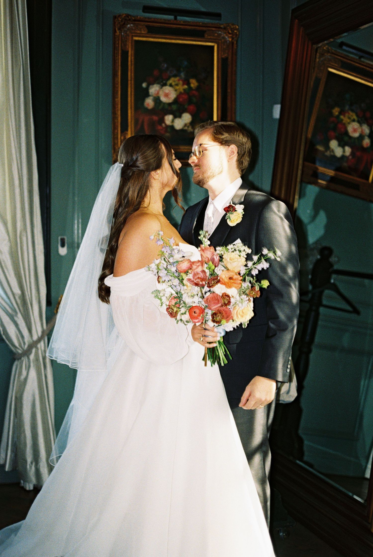 Bride holding colorful bouquet shares tender moment with groom in elegant teal-walled interior.