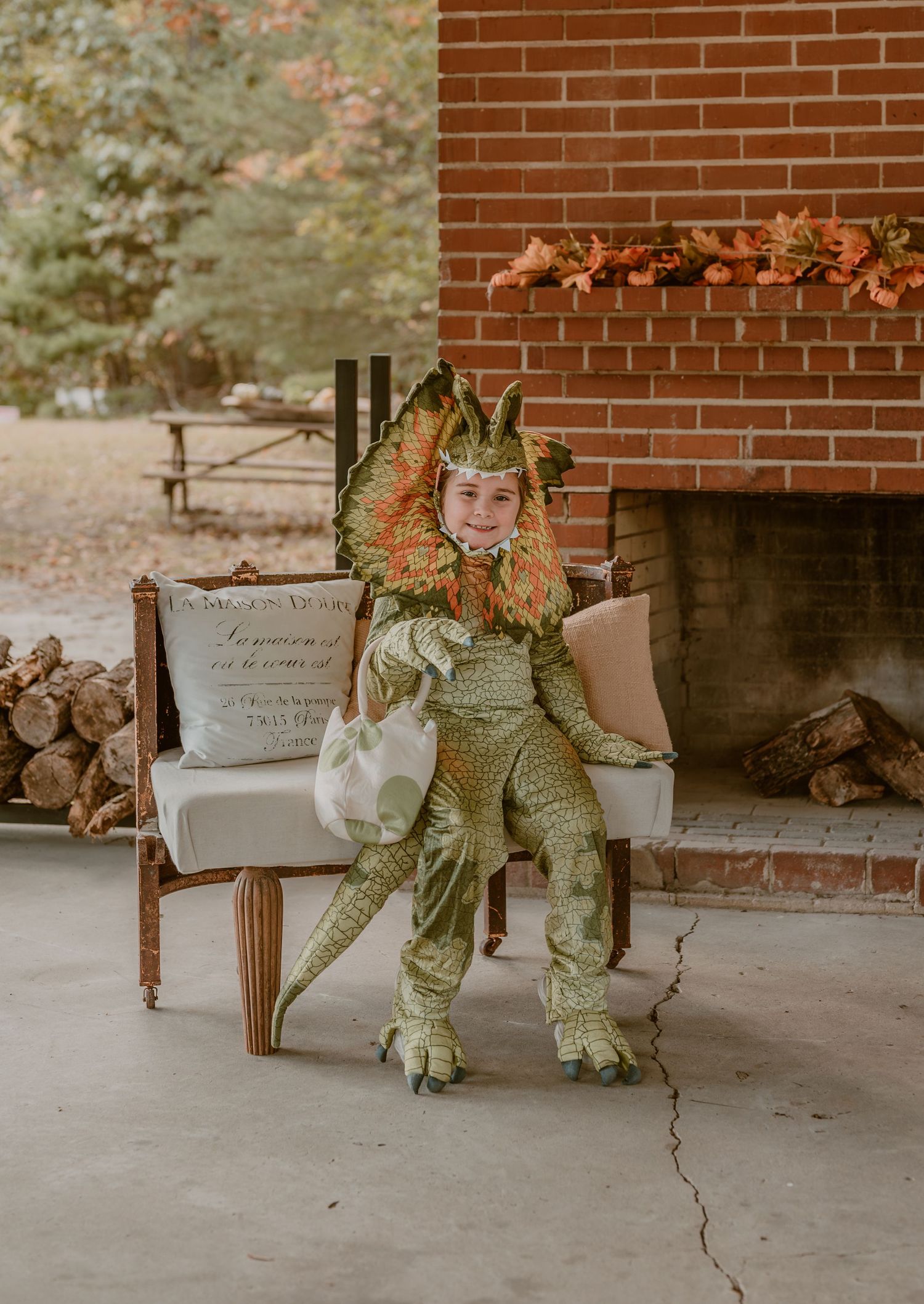 A kid T-Rex dinosaur costume wearing sits in an elegant chair on a patio by a brick fireplace. photo by Samantha Campbell imagery