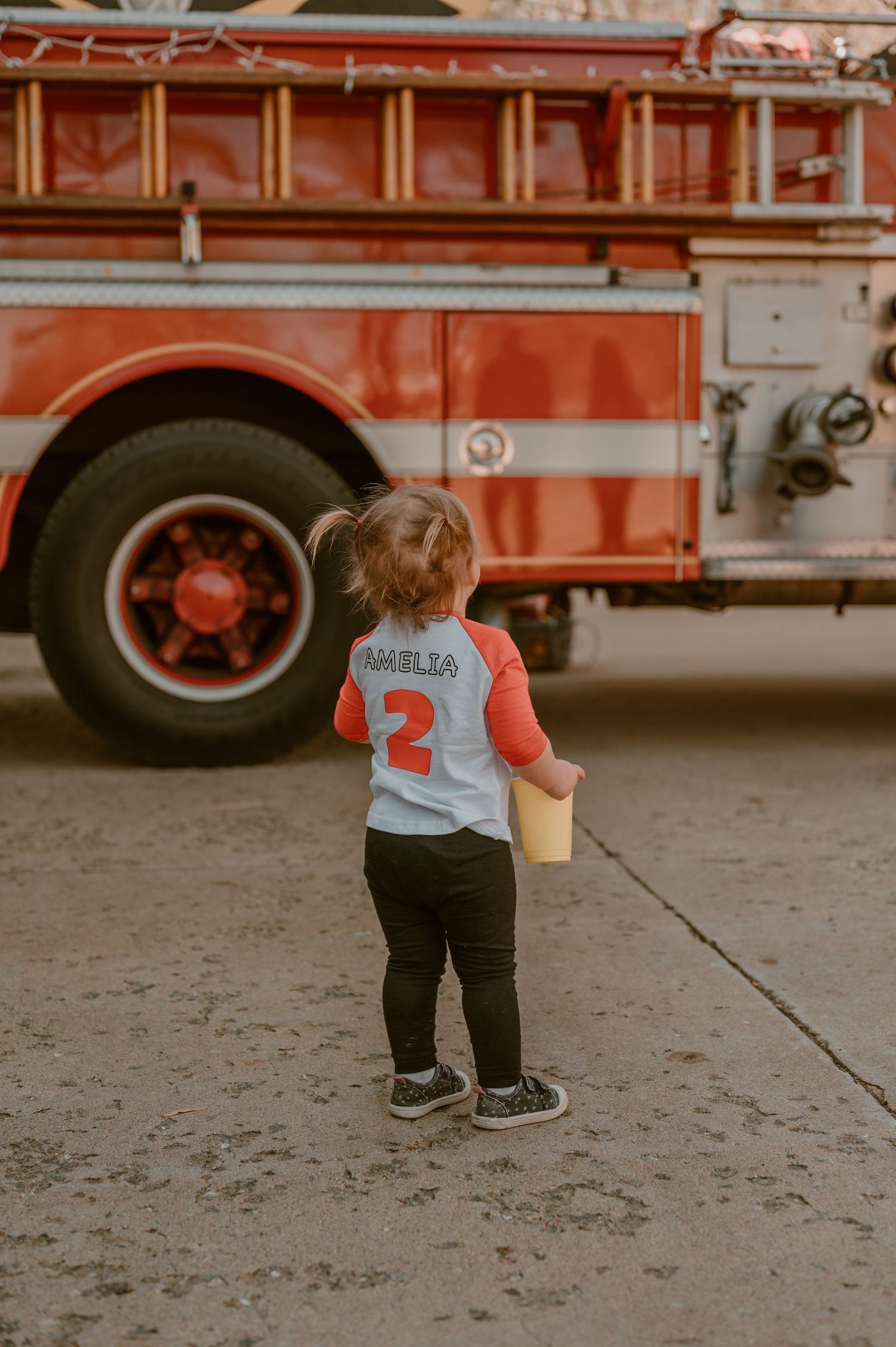 Small child wearing jersey number 2 stands in front of vintage red fire truck on concrete ground. photo by Samantha Campbell imagery