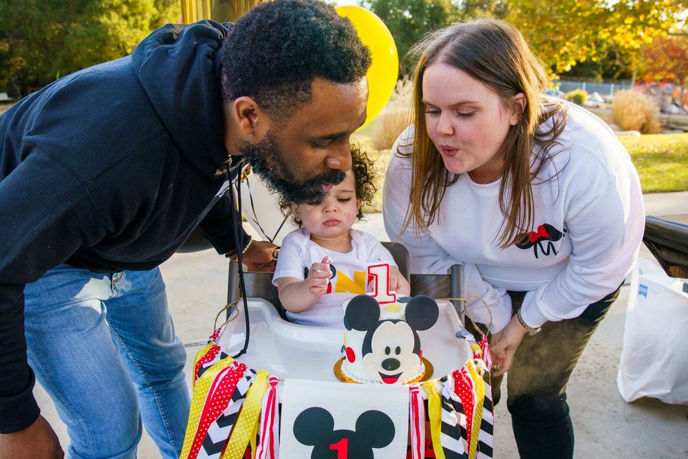 parents helping a young boy blow out his first birthday cake