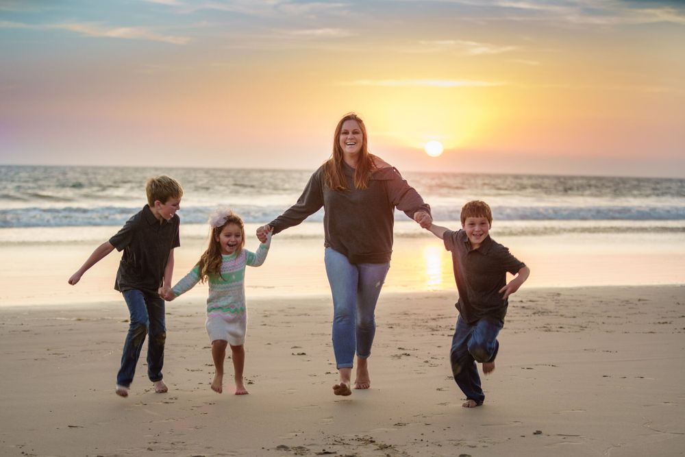 Mom and 3 young kids holding hands, while running on the beach with sunset behind