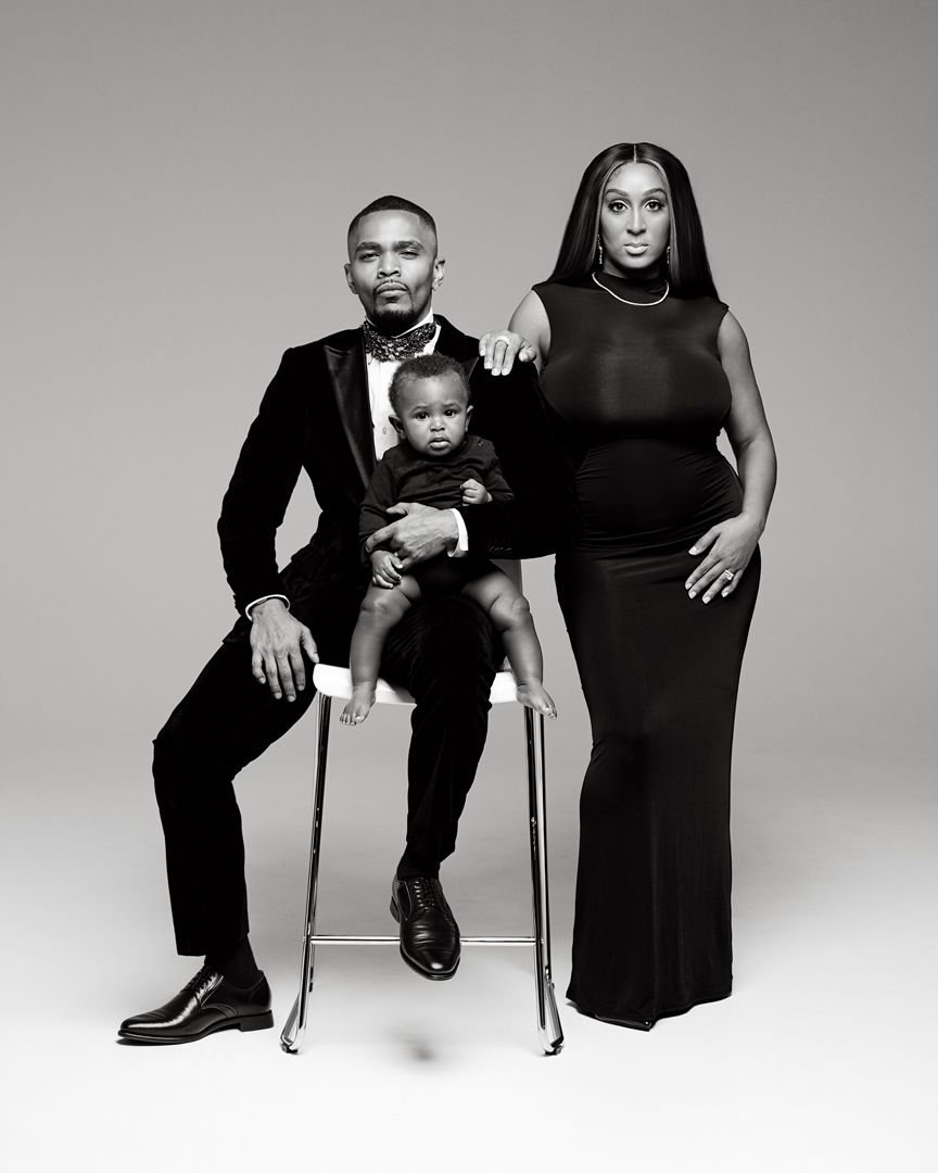 Black and white studio portrait of a family, man in suit seated holding baby, woman in elegant gown standing beside them.