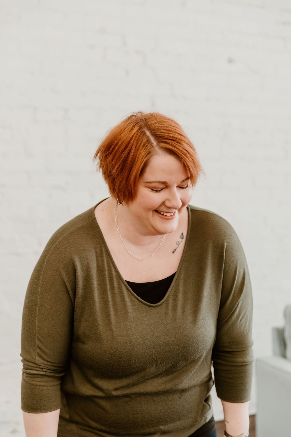A person with short red hair wearing an olive green sweater smiles warmly against a light background.