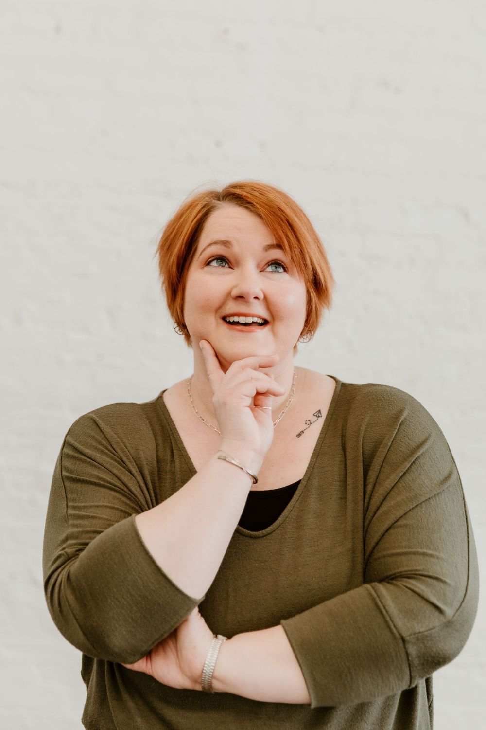 A person with red hair wearing an olive green sweater poses with hand under chin against a light background.