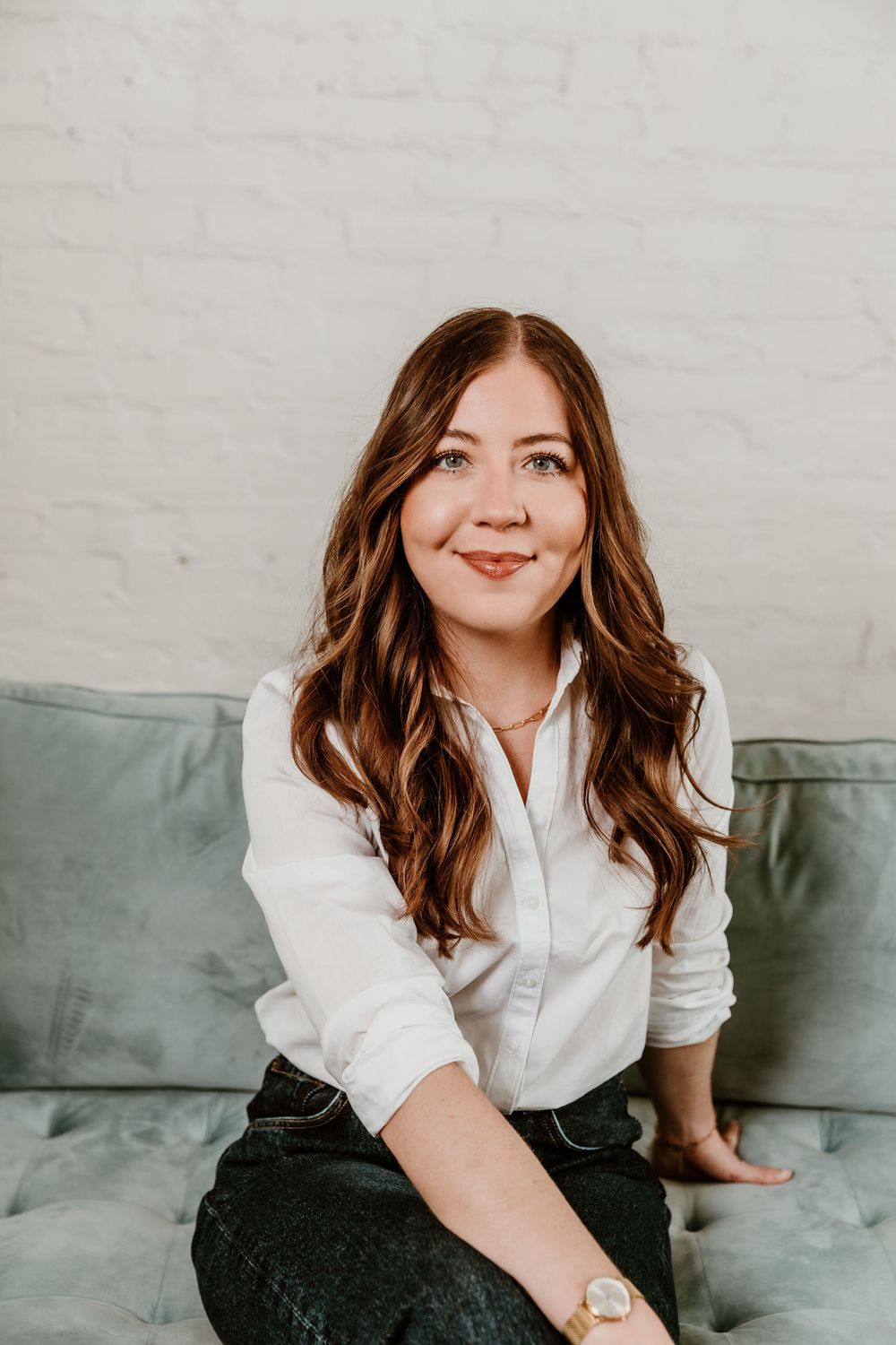 A woman in a white button-down shirt sits casually on a sage green couch with long wavy brown hair.